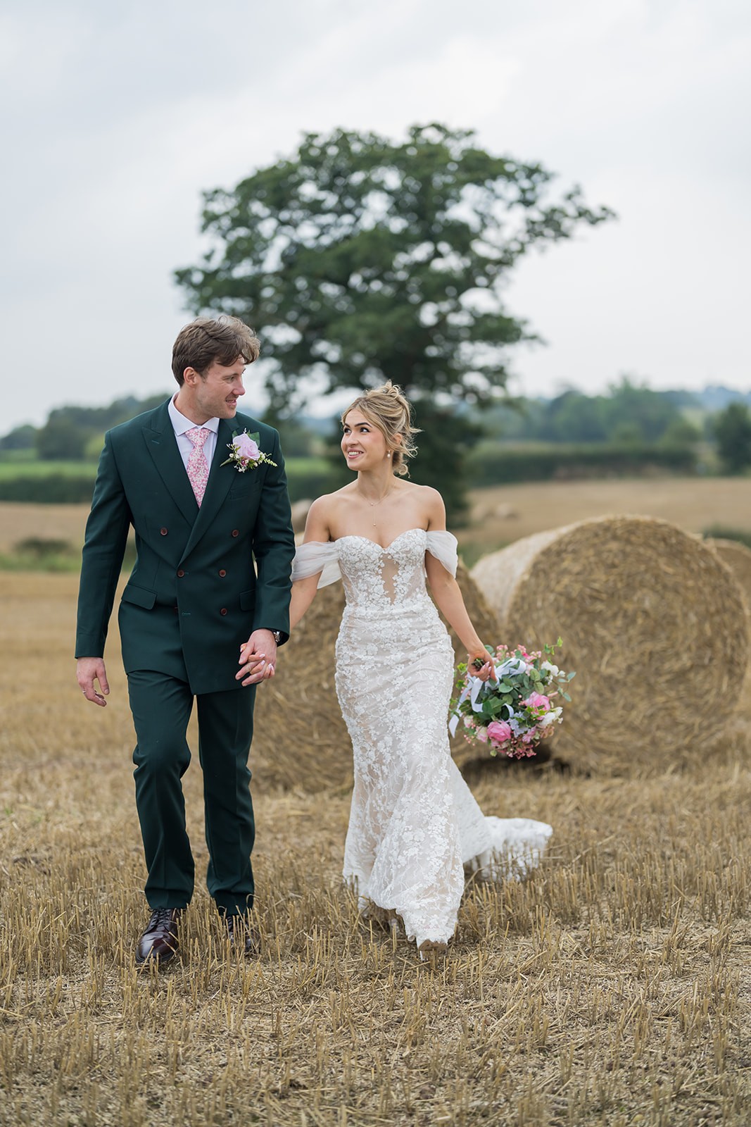 Bride and groom walking through a field beside hay bales